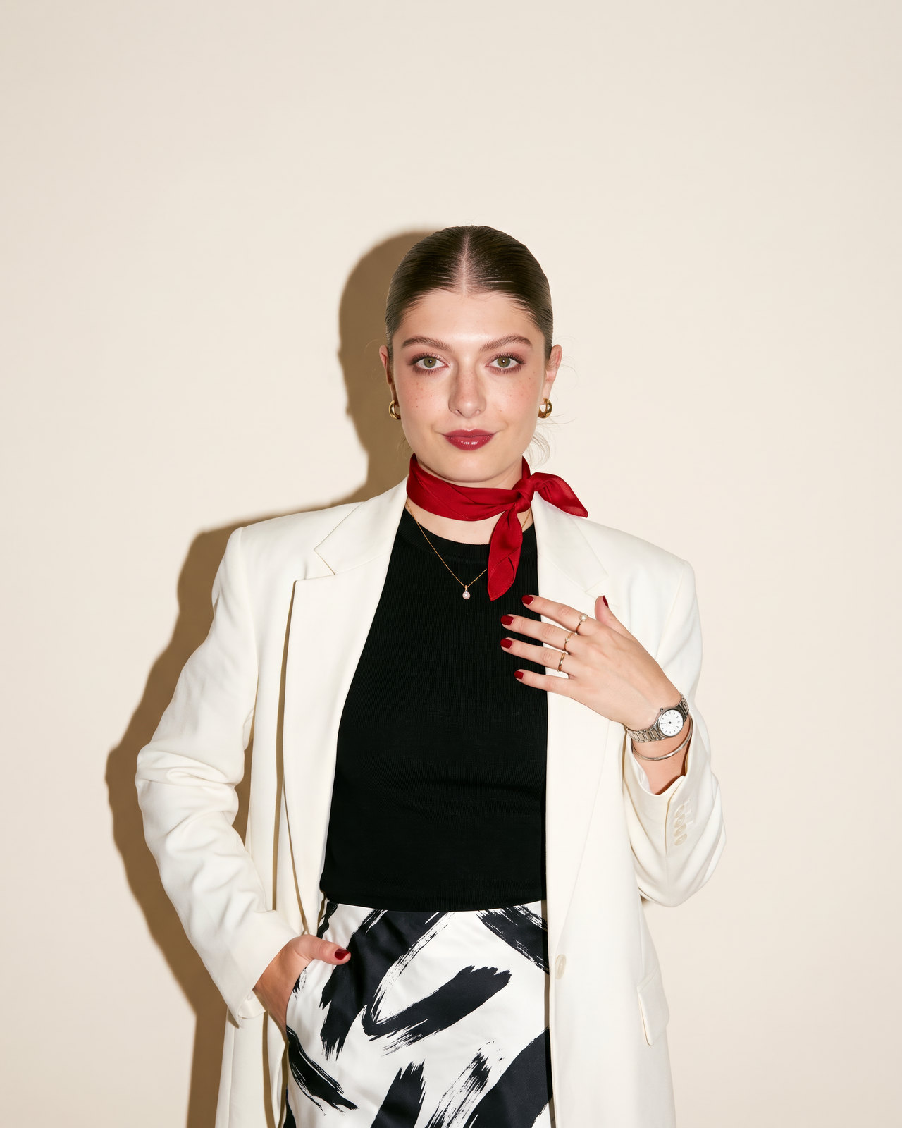 Bela mira a cámara con una mano en el pecho: pelo recogido, blazer blanco, camiseta negra y pañuelo rojo al cuello, con flash directo sobre fondo crema.